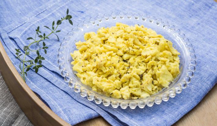 Fotografia em tons de azul e amarelo de uma bandeja de madeira com um pano azul dobrado, sobre ele um ramo de orégano e um pratinho com ovos mexidos.