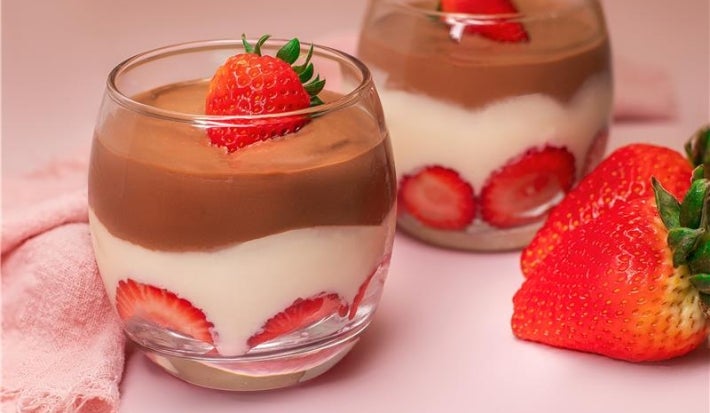 Em um copo transparente, há morangos cortados ao meio, uma camada de creme e uma cobertura de ganache de chocolate.