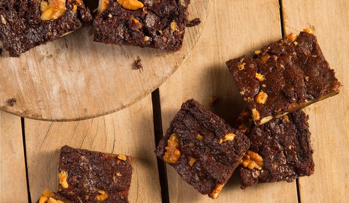 Fotografia em tons de amarelo em uma bancada de madeira com uma tábua de madeira clara com alguns brownies de chocolate em cima. Na bancada, alguns brownies espalhados e flores amarelas espalhadas na mesa para decorar.