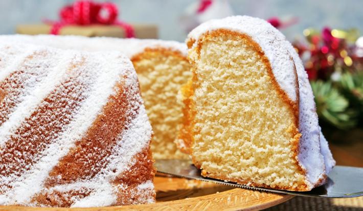 Fotografia de um bolo de massa branca de leite com um pedaço em destaque e cobertura de açúcar de confeiteiro, sobre um apoio reto de tom claro.