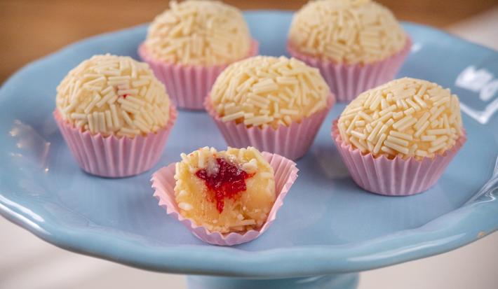 Fotografia de seis forminhas rosa com brigadeiro branco, passado no granulado branco, com recheio de frutas vermelhas. Os brigadeiros estão apoiados em um apoio azul. O brigadeiro da frente está mordido, mostrando a massa e o recheio.