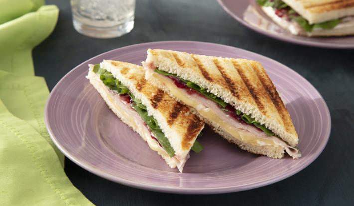 Foto da receita de Panini de Peito de Peru e Queijo Brie. Observa-se um sanduíche cortado na metade sobre um prato de sobremesa lilás recheado.