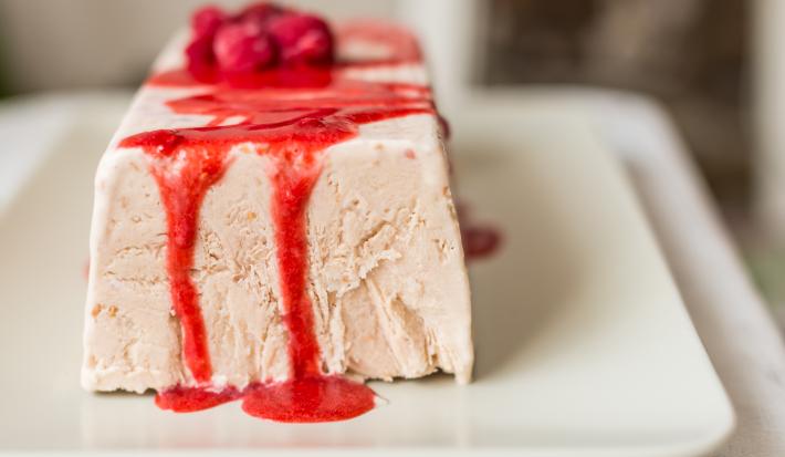 Fotografia de uma terrine de iogurte, em formato de uma forma retangular. Por cima, uma calda de frutas vermelhas, sobre um prato branco quadrangular.