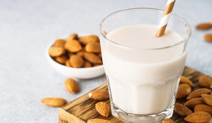Fotografia em tons claros de um copo de vidro com um canudo dourado e branco dentro do copo, que tem uma bebida vegetal de amêndoas. O copo está sobre uma mini tábua de madeira quadrada, apoiada em uma mesa cinza clara com amêndoas espalhadas.