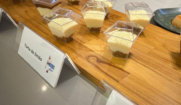 Fotografia em tons de marrom com uma bancada de madeira ao centro. Em cima da bancada existem pequenos potes de sobremesa com uma camada de biscoitos e uma camada de creme branco. A frente desses copos existe uma escrito "Torta de Limão"