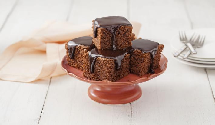 Fotografia em tons de chocolate e branco, ao centro boleira vermelha contendo pedaços de bolo de chocolate com calda de chocolate, ao lado pratos empilhados com 3 garfos por cima, guardanapo rosa, tudo sobre bancada branca.