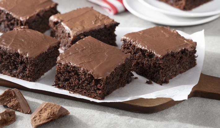 Fotografia em tons de marrom e branco de uma bancada branca  com uma tábua de madeira, sobre ela um papel manteiga com pedaços de bolo de chocolate zero. Ao fundo dois pratos brancos com uma fatia de bolo de chocolate e um paninho vermelho e branco.