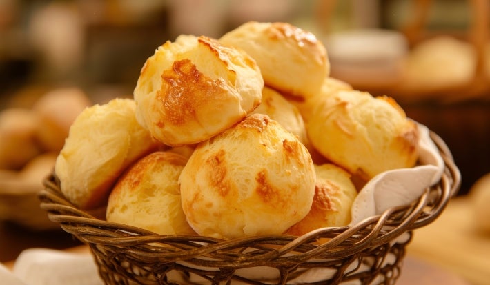 Foto da receita de Pão de Queijos. Observa-se os pães de queijo dispostos numa mesa saindo de uma cestinha de palha.