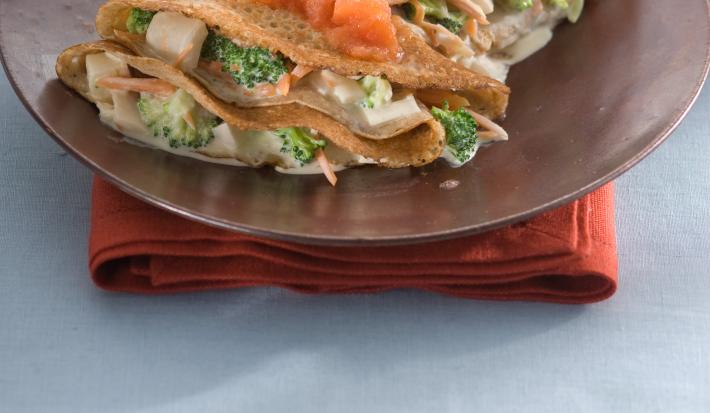 Fotografia de um prato marrom com 2 panquecas feitas em forma de crepe e recheadas com legumes e cobertas com molho de tomate, sobre guardanapo vermelho e toalha azul clara.