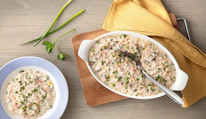 fotografia em tons de cinza e bege de uma bancada cinza vista de cima. Contém uma tábua de madeira com um recipiente oval branco com alças que contém o arroz  com creme, pedaços de carne e ervilhas por cima e ao lado um pano amarelo.