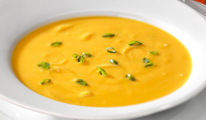 Fotografia em tons de branco e laranja de uma bancada laranja, um supla branco e um prato branco, sobre ele uma sopa de abóbora.