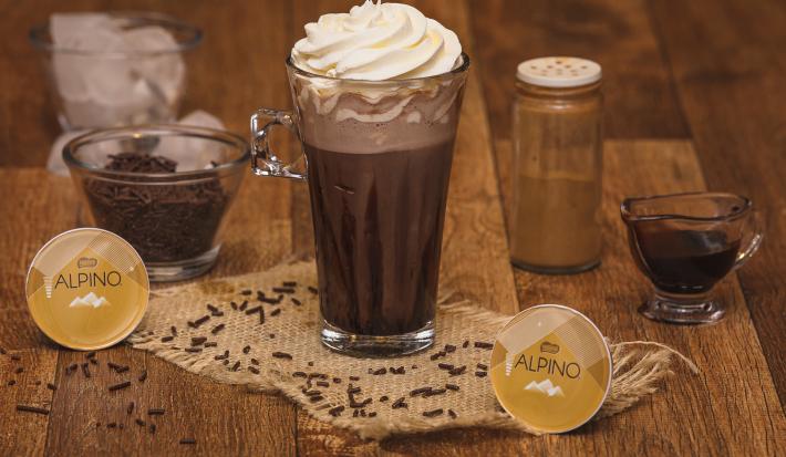 Fotografia em tons de marrom em uma bancada de madeira, com um copo de vidro grande ao centro com a bebida de chocolate Alpino. Ao fundo, um copo com gelo, um potinho com chocolate granulado, e um potinho com canela.