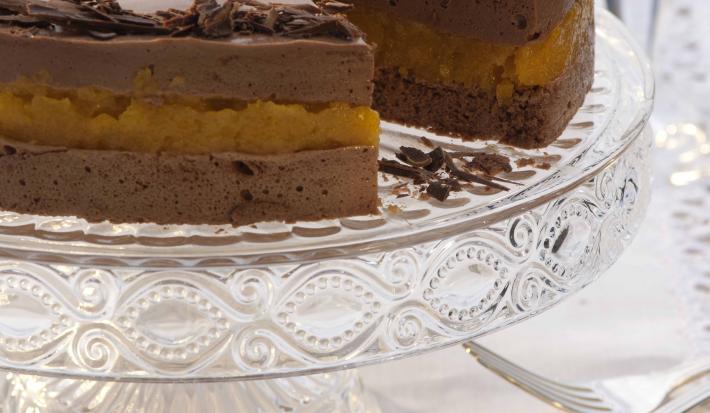 fotografia em tons de branco e marrom de uma bancada com panos branco, contém um suporte para servir sobremesas transparente com uma torta de mousse de chocolate com uma fatia retirada