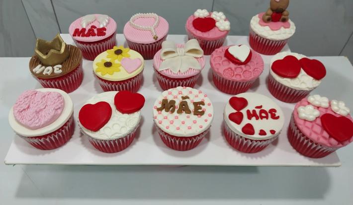 Fotografia em tons de rosa com vários cupcakes ao centro. Cada cupcake possui uma decoração específica, alguns feitos de coração, outros de flores e outros de lacinhos.