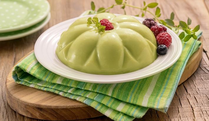 Receita de Manjar com FiberMais Suco Verde. Na foto, observa-se um manjar esverdeado sobre um prato branco que está em cima de um guardanapo em tons de verde e uma tábua oval de madeira. Frutas vermelhas e um ramo de hortelã decoram a sobremesa.