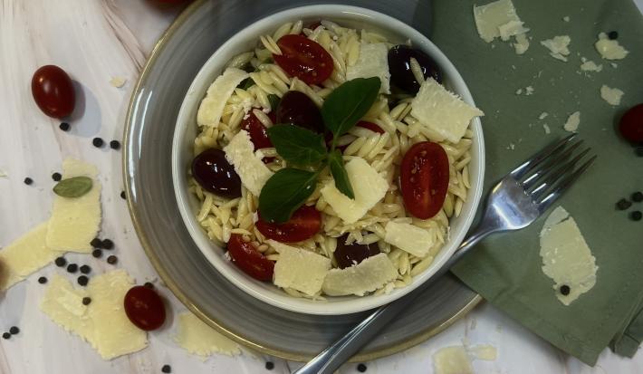 Foto da receita da salada de risoni servida em um bowl branco em cima de um prato de vidro transparente com um garfo prateado ao lado e um pano verde decorativo. O prato em cima de uma mesa clara de madeira com os ingredientes da salada como decoração.