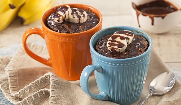 Fotografia em tons de beje, azul e laranja de uma bancada branca com um paninho beje, sobre ele um caneca azul e uma caneca laranja com bolo de caneca. Ao fundo um cacho de banana e um recipiente redondo com calda.