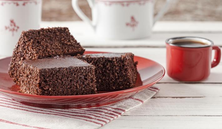 Fotografia em tons de vermelho em uma bancada de madeira clara com um pano listrado em branco e vermelho e um prato vermelho com três pedaços de bolo de cacau com coco. Ao lado, uma xícara vermelha de café e ao fundo, dois bules brancos.