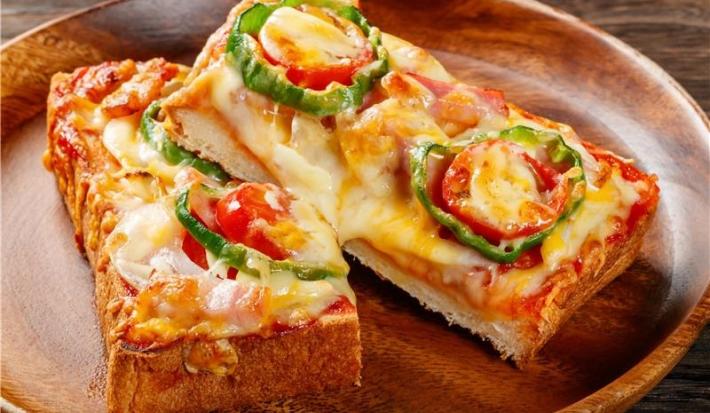 Fotografia de um pão pizza de queijo deretido com tomate, cortado na metade e a outra metade esta apoiado sobre a outra metade. As metades estão em um prato redondo de madeira raso.