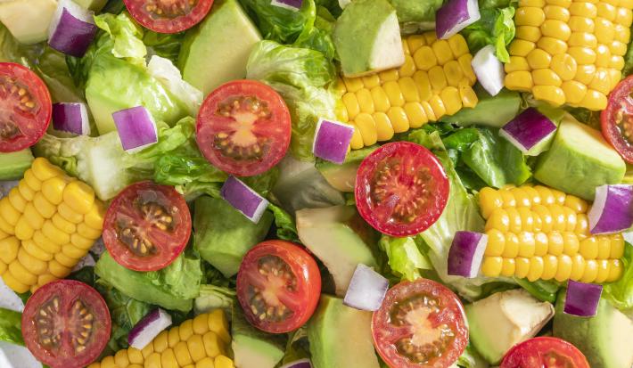 Fotografia vista de cima de uma salada mista com pequenos pedaços da espiga de milho, tomates cereja, cebola roxa, alface e chuchu. A salada está dentro de um recipiente de vidro branco, sobre uma toalha de mesa branca. Ao redor, tomates, abacate e limão.