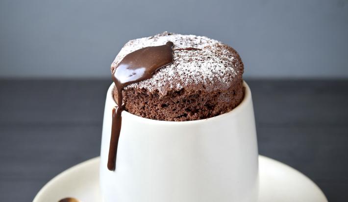 Fotografia de um suflê de café com NESCAFÉ e açúcar de confeiteiro polvilhado por cima. A sobremesa está em um recipiente de vidro branco alto em cima de um prato pequeno branco de sobremesa, e uma colher de café apoiada.