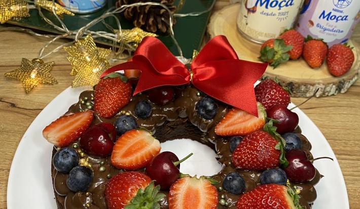 Foto da receita de guirlanda de brownie decorada com frutas vermelhas e decoração de natal servida em um prato grande branco sobre uma mesa de madeira com produtos de leite moça ao fundo