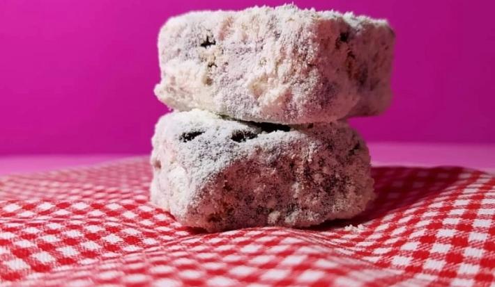 Foto da receita de Palha Italiana. Observa-se duas palhas italianas cortadas em quadrados dispostas em uma toalha quadriculada branca e vermelha e em um fundo rosa.
