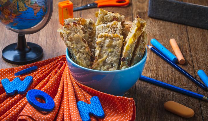 Fotografia em tons de laranja e azul em uma bancada de madeira escura, um paninho laranja e vários objetos escolares na cena. Ao centro, um potinho azul com várias barrinhas de cereal com banana e Mucilon dentro dele.
