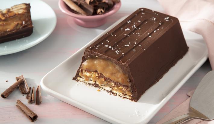 Foto da receita de Barra de chocolate e caramelo crocante. Observa-se uma barra de chocolate cortada em uma travessa grande. Ao lado direito, uma espátula de cortar e, ao esquerdo, um pedaço da torta cortada