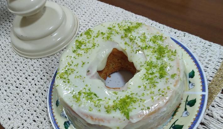 Foto em tons de branco da receita de bolo de nescafé com cobertura de limão servida em uma porção grande sobre um prato decorado em cima de uma mesa de madeira com um pano bordado