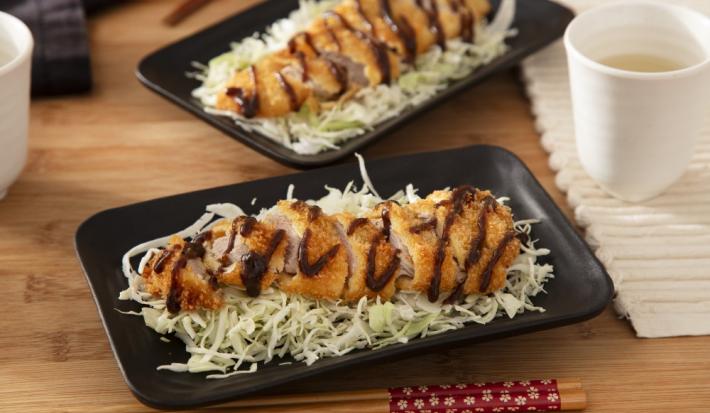 Foto da receita de Tonkatsu (Bife de Porco Japonês). Observa-se dois pratos retangulares pretos com repolho fatiado e o tonkatsu por cima, servido com um pouco de molho escuto. Observa-se um hashi na foto, na frente do prato.