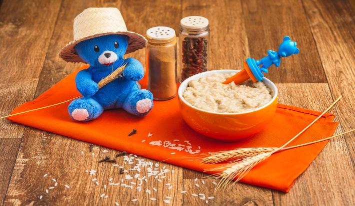 Fotografia em tons de laranja e azul de uma bancada de madeira com um paninho laranja e um pote laranja com a papinha. Na frente, grãos de arroz e cravos decorando e uma pelúcia azul com chapéu de palha.