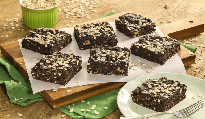 Fotografia em tons de marrom e verde de uma bancada de madeira com um paninho verde com uma tábua de madeira, sobre ela um papel manteiga com fatias de brownie. Ao lado um prato redondo verde com um brownie e um garfo. Ao fundo um pote com aveia.