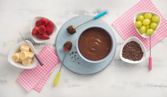fotografia em tons de rosa e cinza de uma bancada vista de cima, ao centro panos listrados na cor rosa e branco, uma mini tabua azul com um recipiente redondo em cima com chocolate, 4 potes brancos um com granulado e os demais com frutas.