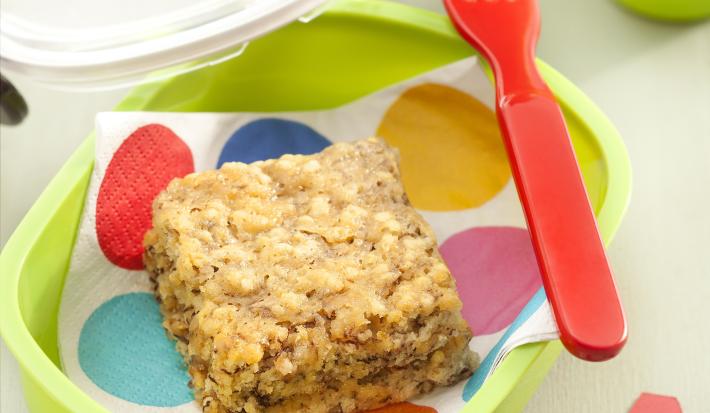 Fotografia em tons de verde em uma bancada cinza ao centro um potinho verde com um guardanapo de bolas coloridas e um pedaço do bolo de banana e quinoa, ao lado um garfo vermelho.