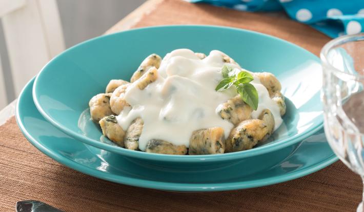 Fotografia em tons de azul em uma bancada de madeira, um paninho azul com bolinhas brancas, prato redondo verde com o nhoque nutritivo com molho branco.