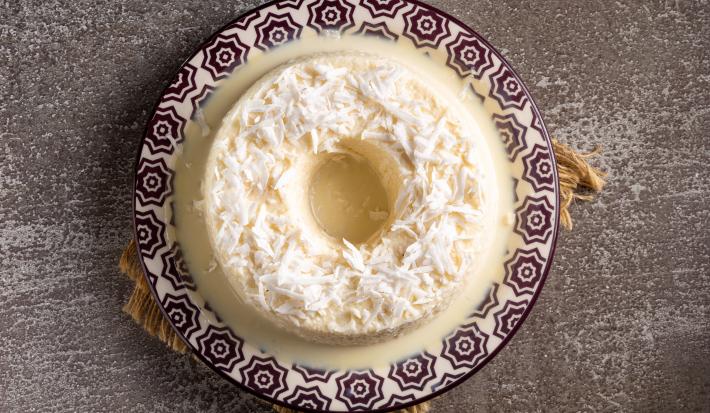 Fotografia vista de cima de um prato branco com desenhos de tom escuro com um pudim de coco com raspas de coco por cima. O prato está apoiado sobre um pano marrom com rebarbas decorativas.