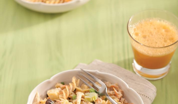 Fotografia em tons de verde em uma bancada de madeira verde com um pano bege, um prato fundo bege com a salada colorida com macarrão, ao lado um copo de vidro com um suco de laranja.