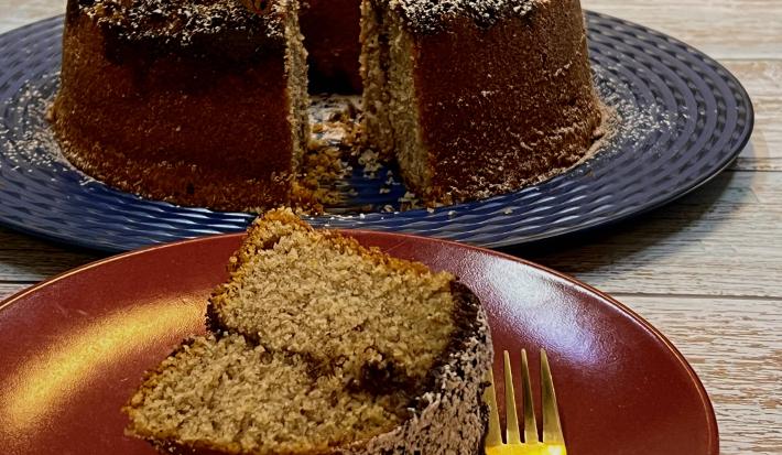 Foto da receita de Bolo com Farofa de Canela. Observa-se um prato de sobremesa com uma fatia do bolo e um garfinho e, atrás, o bolo maior em um prato grande azul.