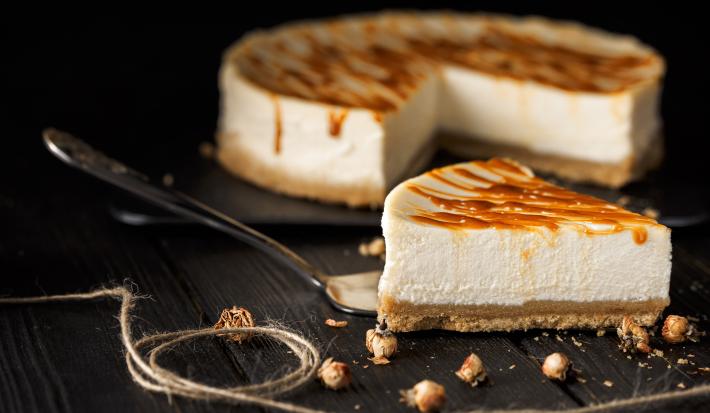 Fotografia de uma fatia de cheesecake com caramelo salgado por cima de uma espátula de prata, sobre uma mesa preta. No fundo tem o resto da cheesecake sobre um apoio preto.