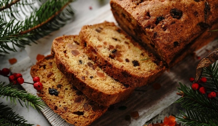 Fotografia de um bolo de frutas cortado, exibindo sua textura densa e rica, com pedaços de frutas secas e nozes. O bolo está disposto sobre uma superfície rústica, cercado por decorações de ramos verdes e bagas vermelhas.