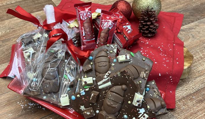 Fotografia em tons vermelhos e marrons em uma bancada de madeira marrom claro. Ao centro um prato quadrado vermelho com a receita da barra de chocolate quebrada em pedaços sendo uma exposta e outra na embalagem para presente sobre um tecido vermelho