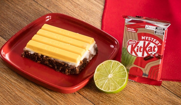 Fotografia em tons de vermelho com uma fatia de brownie ao centro. Por cima do brownie existe um glacê de limão coberto com 4 pedaços de kitkat Mystery, o kitkat de sabor misterioso de cor amarela. Fotografia em tons de vermelho com uma fatia de brownie ao centro. Por cima do brownie existe um glacê de limão coberto com 4 pedaços de kitkat Mystery, o kitkat de sabor misterioso de cor amarela.