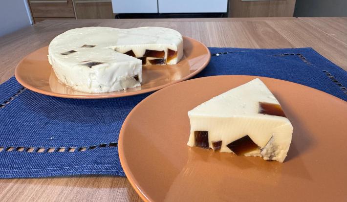 Fotografia de dois pratos rasos de tom marrom claro apoiados em uma mesa de madeira com um pano azul quadrado. Sobre o prato da frente tem uma fatia grande de gelatina cremosa de leite moça com cubinhos de gelatina de café. No outro tem o restante.