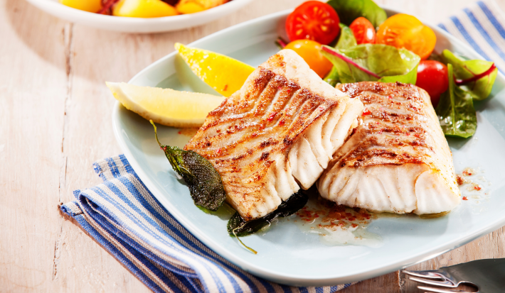 Fotografia mostra dois filés de peixe grelhado sobre um prato branco acompanhado de salada.