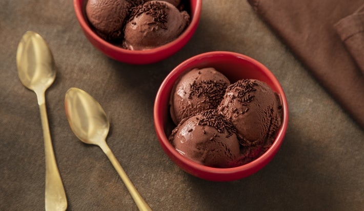 Fotografia de dois recipientes de vidro fundos, pequenos e vermelhos, ao lado de duas colheres de sopa douradas. Dentro dos recipientes têm três bolas de sorvete de nescau extra cacau com granulado por cima. Fotografia de dois recipientes de vidro fundos, pequenos e vermelhos, ao lado de duas colheres de sopa douradas. Dentro dos recipientes têm três bolas de sorvete de nescau extra cacau com granulado por cima.