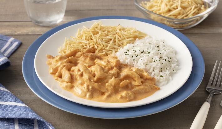 Fotografia em tons de azul em uma bancada de madeira com um sousplat azul, um prato branco raso em cima com o strogonoff, arroz branco e batata palha. Ao lado, um pano azul com listras brancas, talheres, copo com água e um potinho com batata palha.