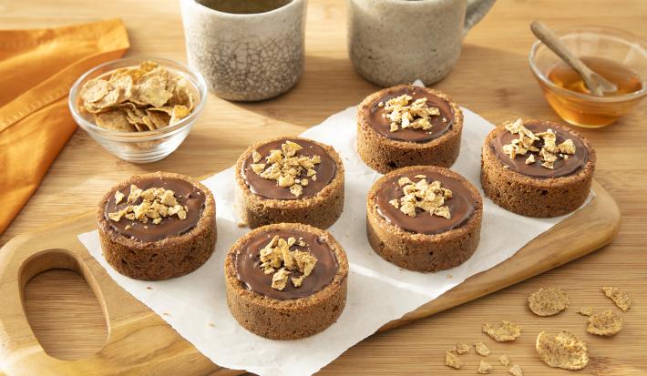 Foto da Receita de Torta de Banana e Cacau com Nesfit Cereal. Observa-se uma tábua de madeira com 6 tortinhas sobre ela, recheadas de chocolate e polvilhas de cereal nesfit por cima. Cereais e um potinho com mel decoram a foto.