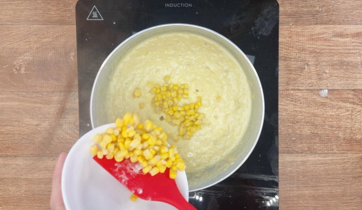 Creme de milho cozinhando em panela sobre fogão portátil preto. Uma espátula despeja milho verde cozido de uma tigela dentro da panela