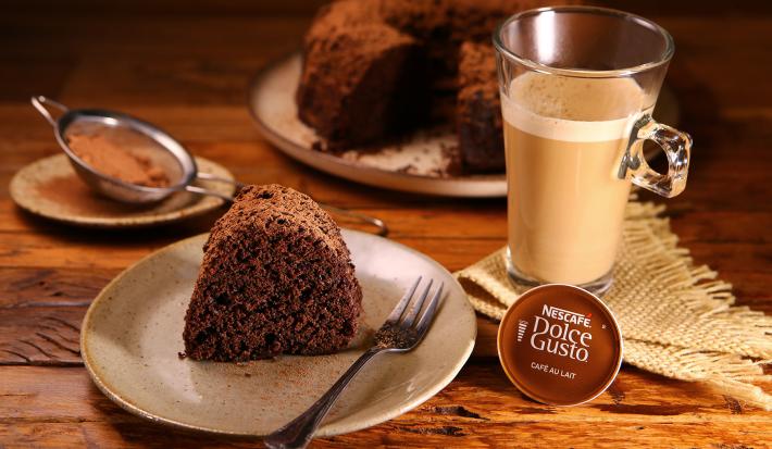 Fotografia em tons de marrom em uma bancada de madeira clara, um guardanapo de pano bege de palha, uma xícara em cima com o Café ao Leite Dolce Gusto. Ao centro, um prato redondo com um pedaço do bolo de chocolate em cima dele. Ao fundo, o bolo inteiro.